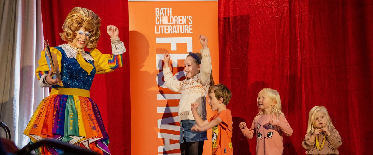 Drag queen Mama G reading a book to a group of children. They all have their hands in the air and are smiling