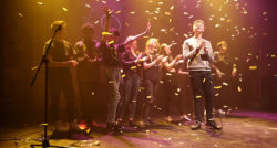 A group of young people onstage clapping as confetti falls from the ceiling