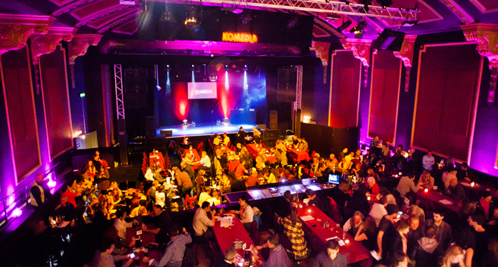 Komedia is full with lots of tables of people in front of the stage. Everything is lit up in multicoloured lights.