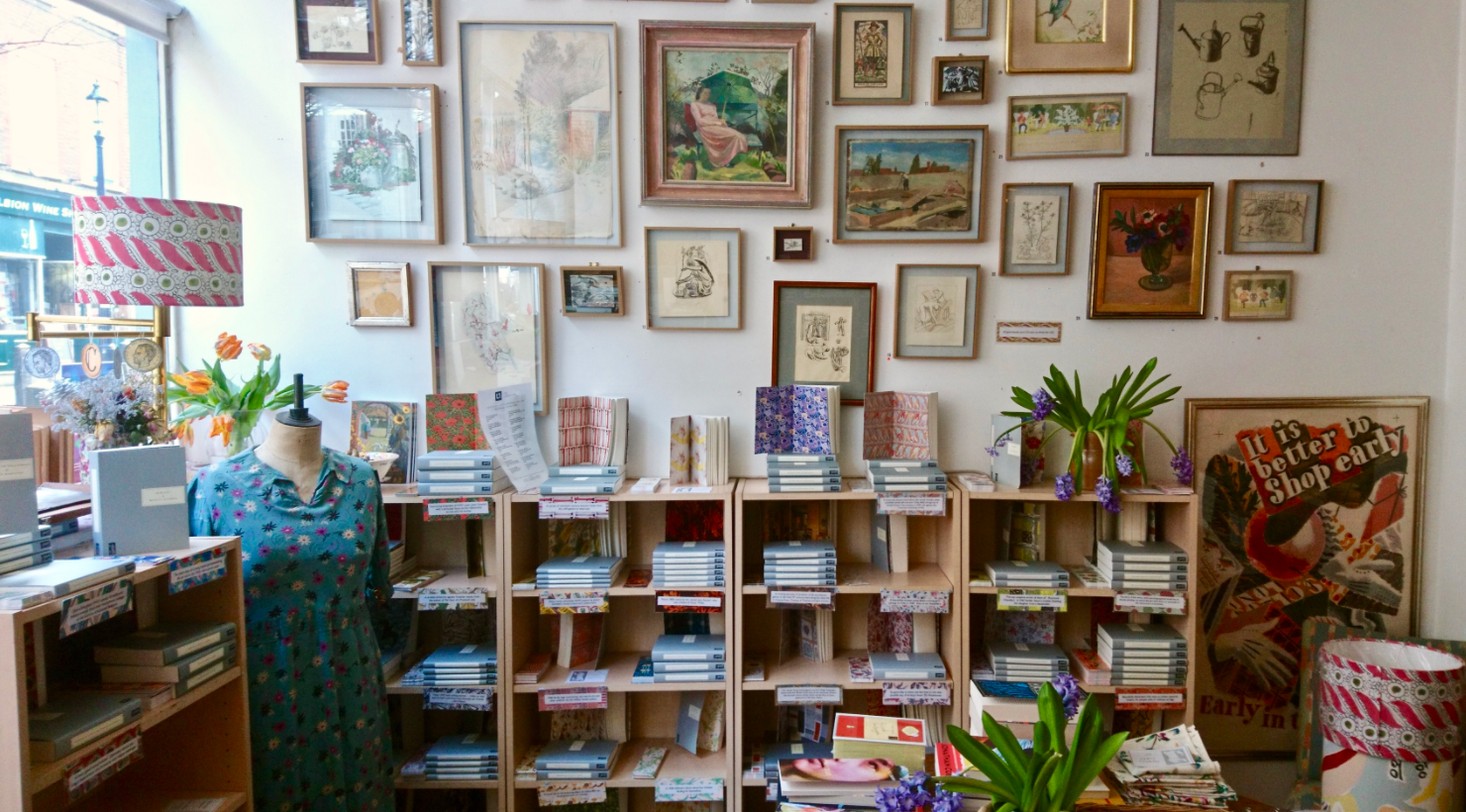 Inside Persephone Bookshop – the wall is covered in bookshelves with lots of paintings hanging above them